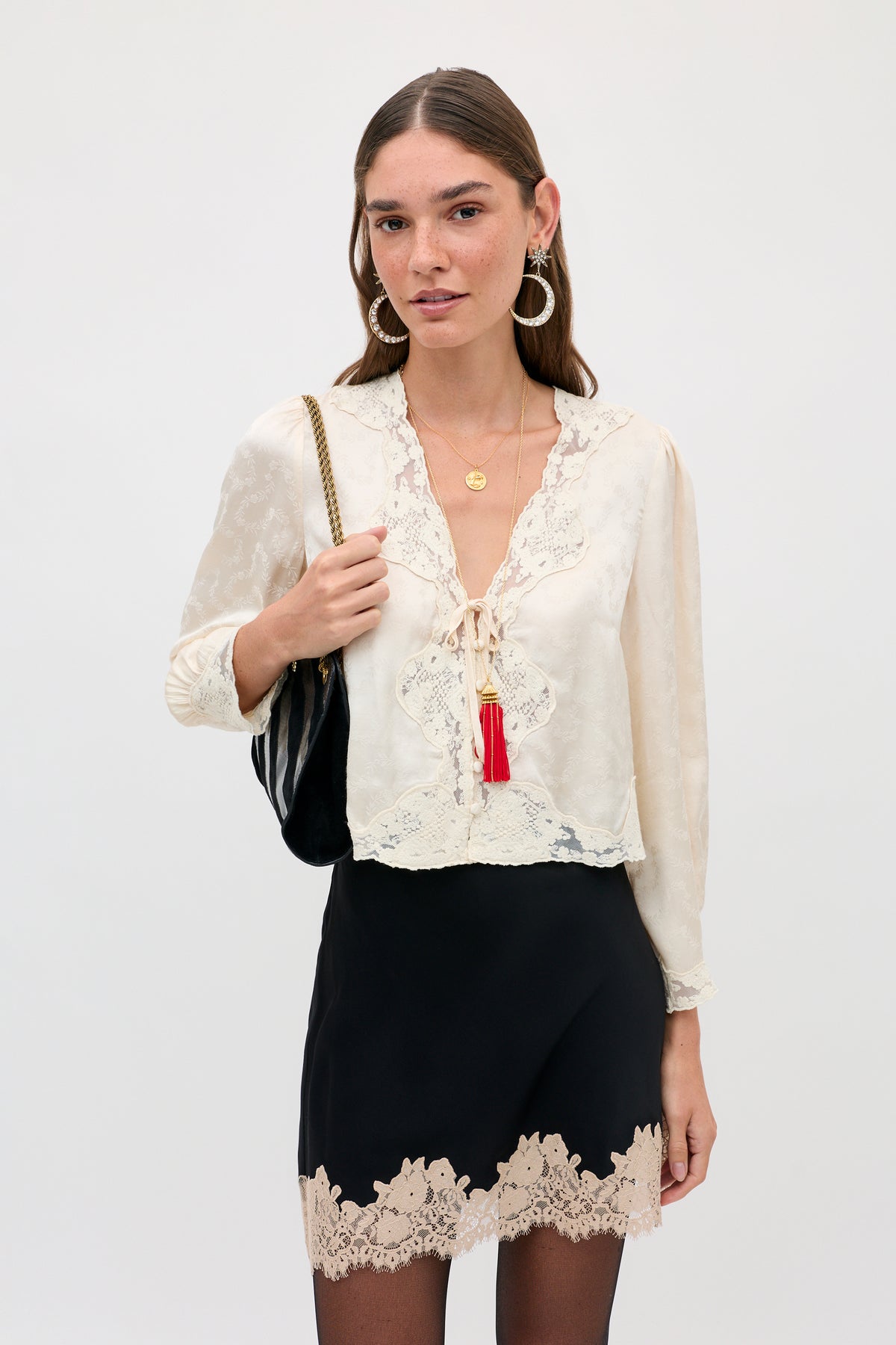 A woman wears a cream-colored long-sleeved blouse with lace trim and a red tassel, paired with an Ariah - Black skirt featuring a lace hem and a black shoulder bag.