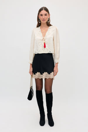 A model wears the Ariah - Black blouse, a long-sleeved cream top with lace trim and a red tassel tie, paired with a black skirt featuring a lace hem, black tights, black knee-high boots, and a small black handbag.