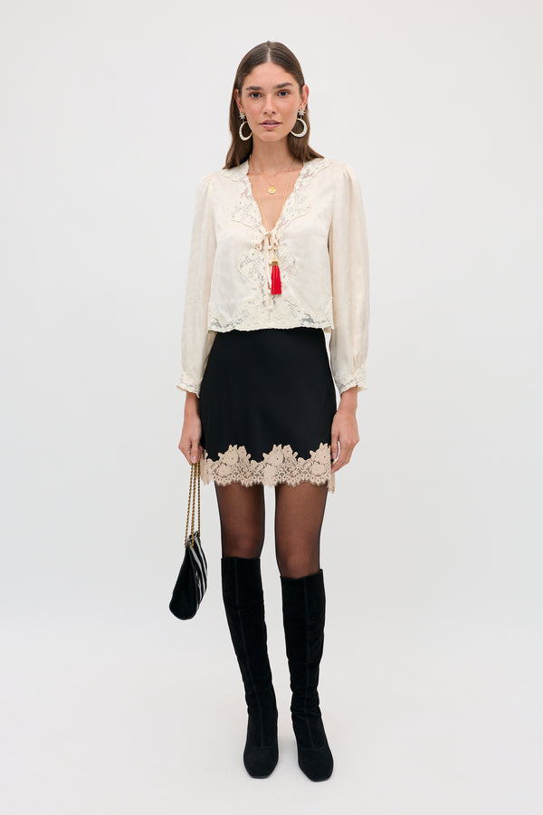 A model wears the Ariah - Black blouse, a long-sleeved cream top with lace trim and a red tassel tie, paired with a black skirt featuring a lace hem, black tights, black knee-high boots, and a small black handbag.
