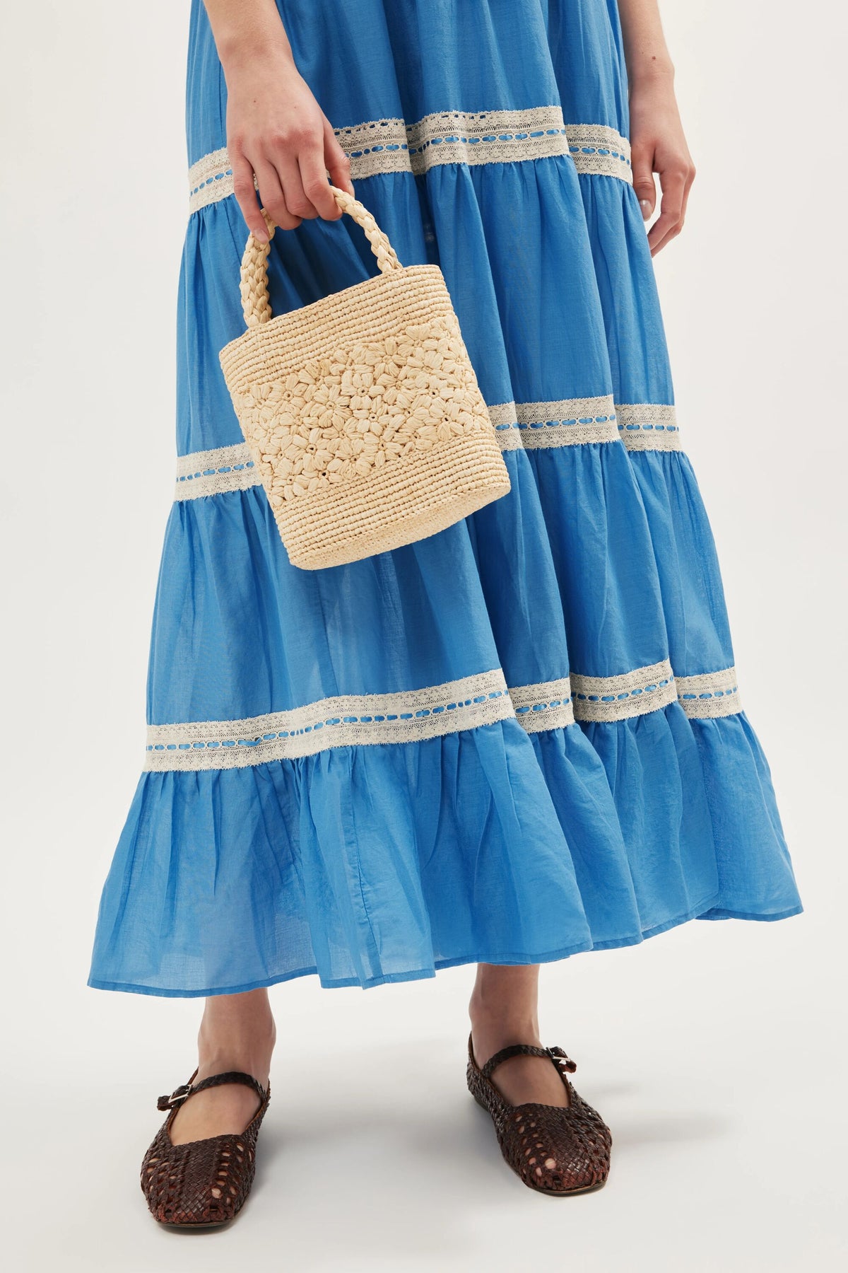 A person wearing the Aubrie - Blue dress, featuring lace trim and delicate shirred panels, holds a small woven straw bag and wears brown woven flat shoes.