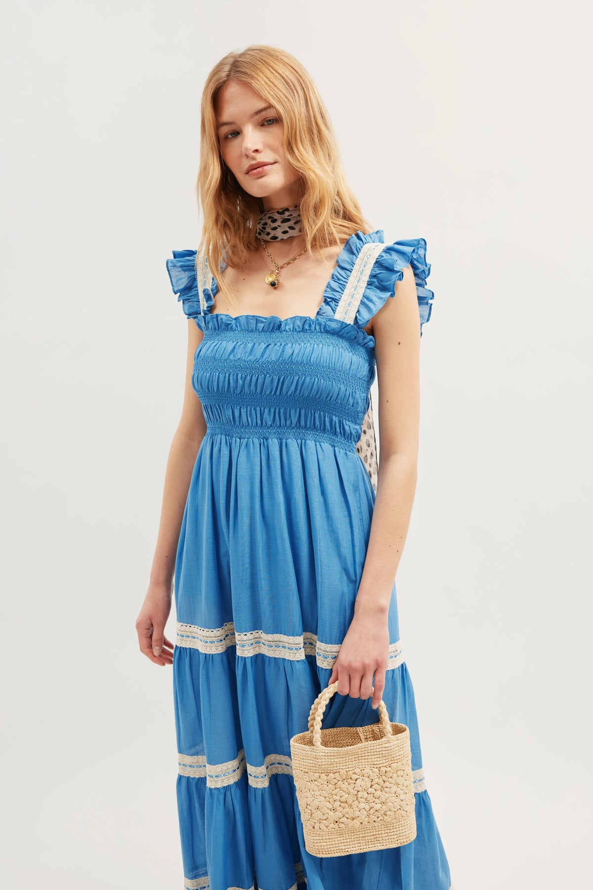 A woman wearing the Aubrie - Blue dress, featuring blue ruffles, sleeveless design, white lace trim, and delicate shirred panels, holds a small woven basket and wears a patterned scarf around her neck.