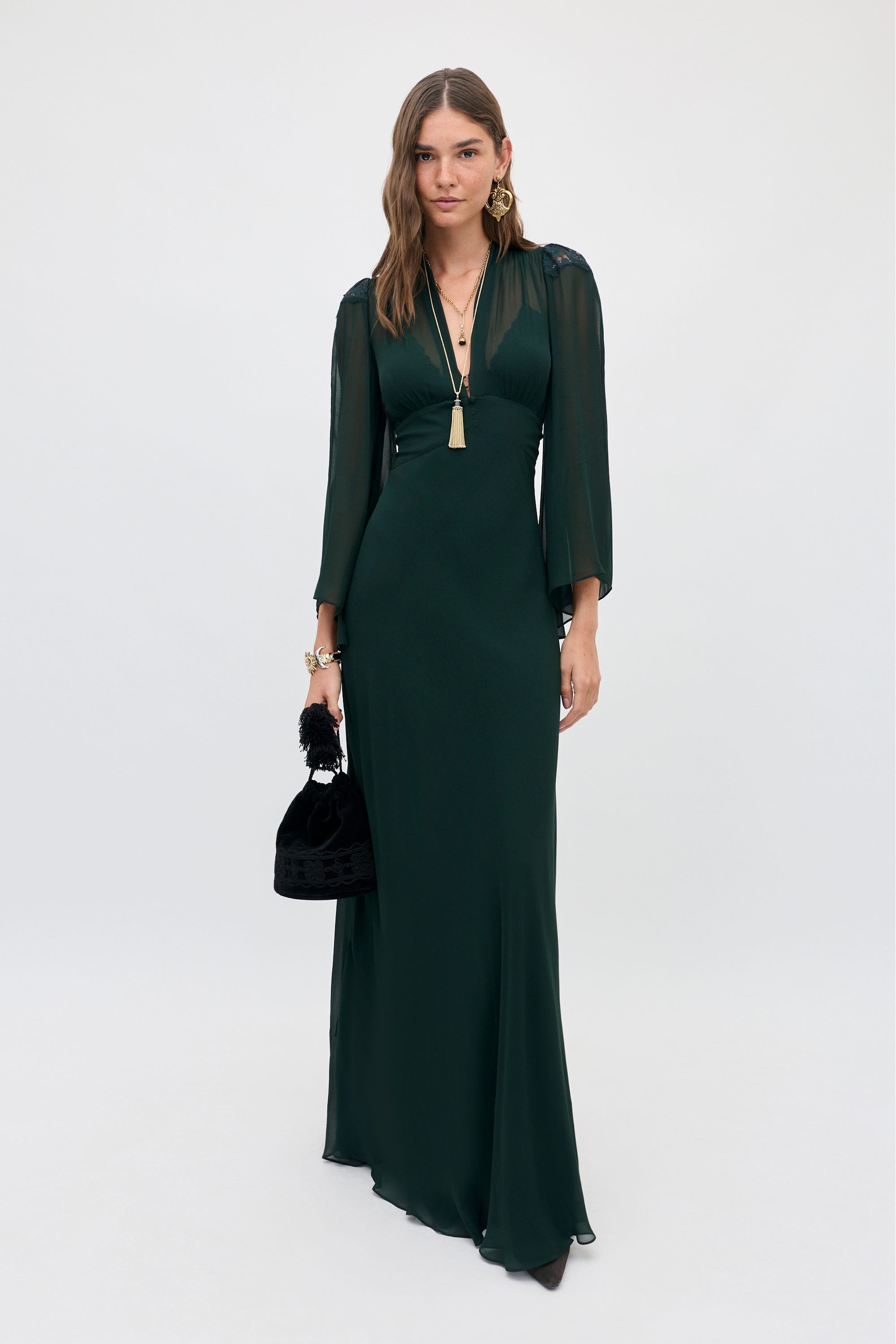 A woman wears a Biba - Dark Green gown with a V-neck, sheer long sleeves, and a small black handbag.