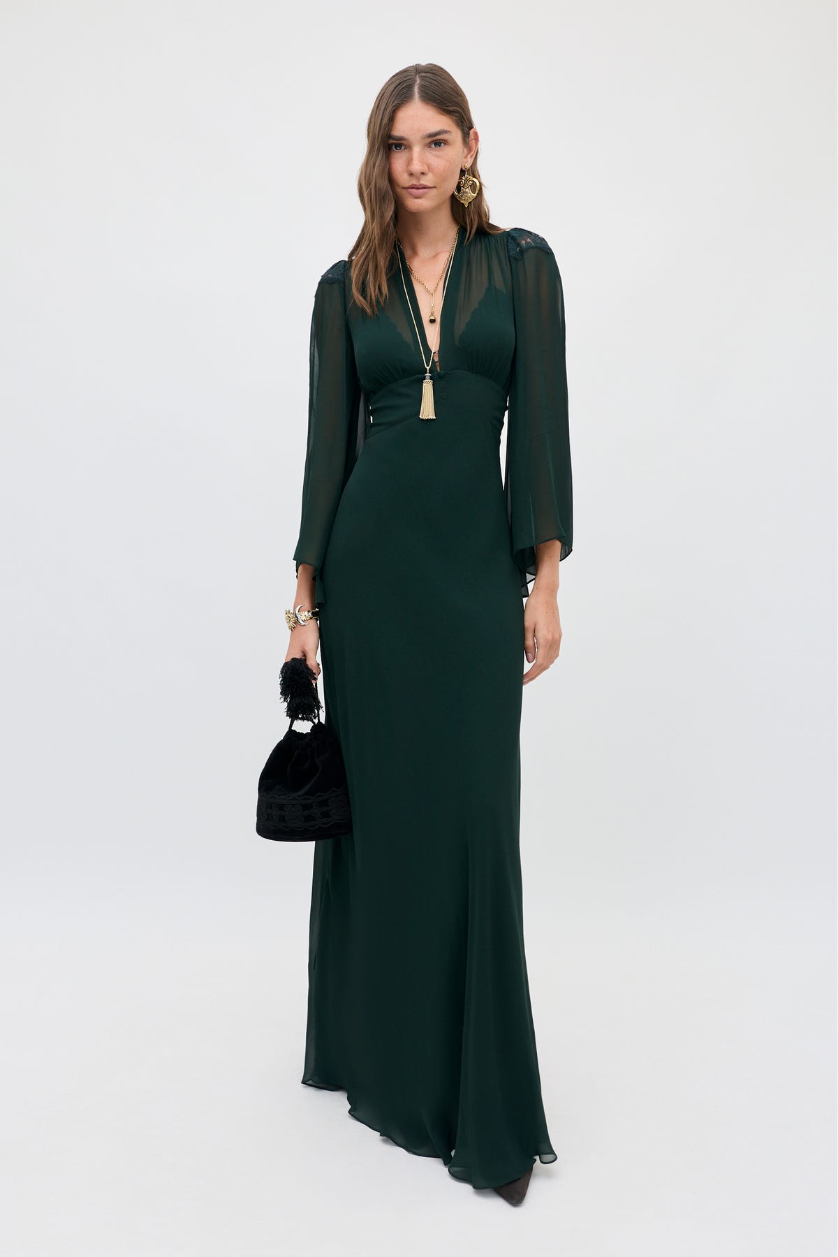 A woman wears a Biba - Dark Green gown with a V-neck, sheer long sleeves, and a small black handbag.