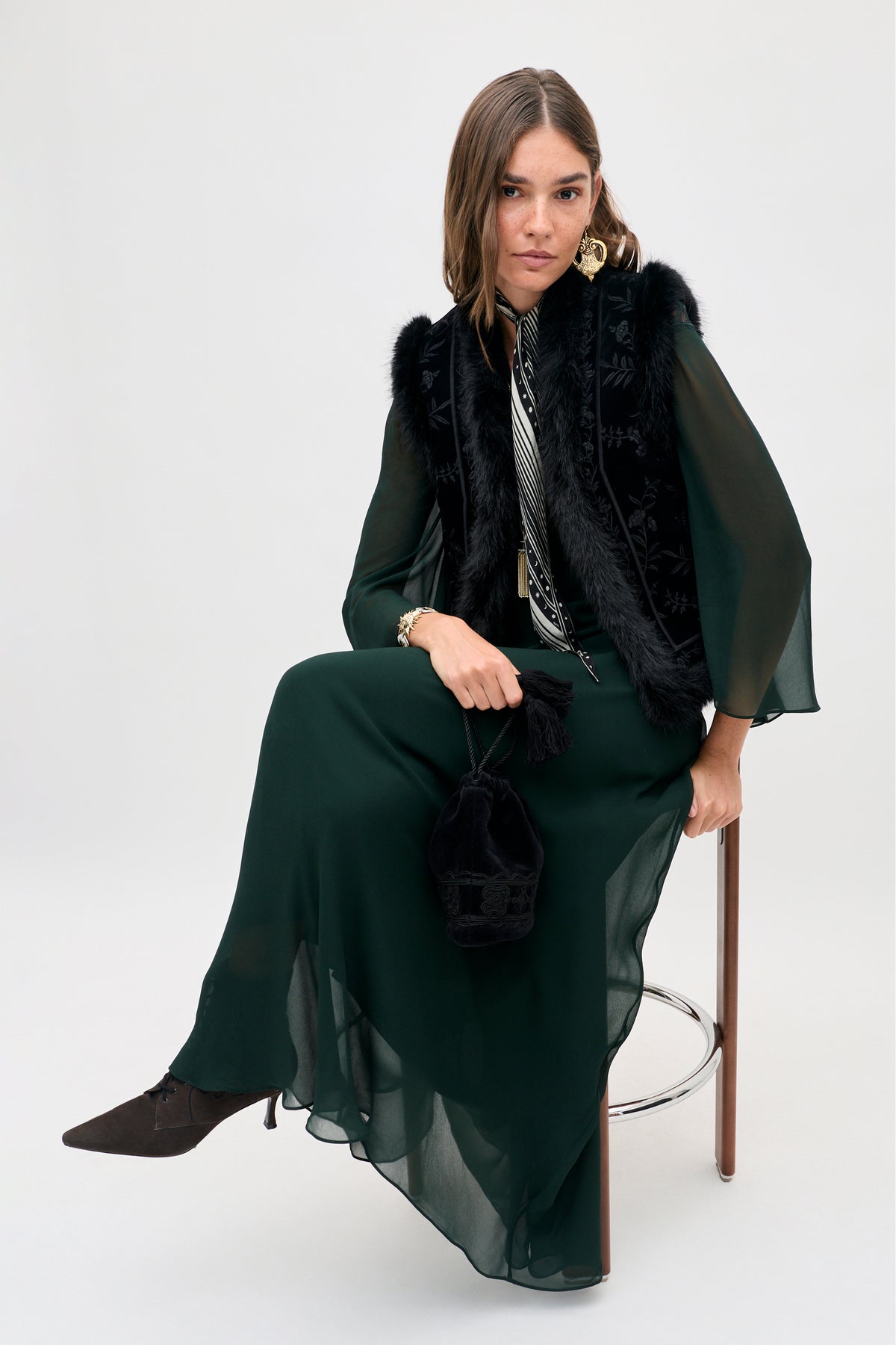 A woman wearing a dark green long-sleeved dress and a Taletha - Botanical Vine Embroidery Black vest with fur trim, sitting on a stool and holding a small black drawstring bag.