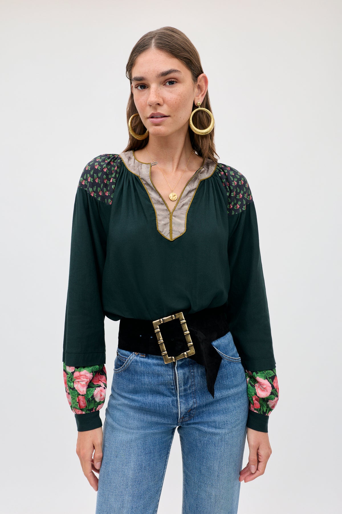 A woman wears the Bria - Rose Mix Pink dark green long-sleeved blouse with floral patterns on the shoulders and cuffs, paired with a black belt featuring a large gold buckle and blue jeans.
