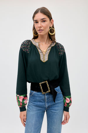 A woman wears the Bria - Rose Mix Pink dark green long-sleeved blouse with floral patterns on the shoulders and cuffs, paired with a black belt featuring a large gold buckle and blue jeans.