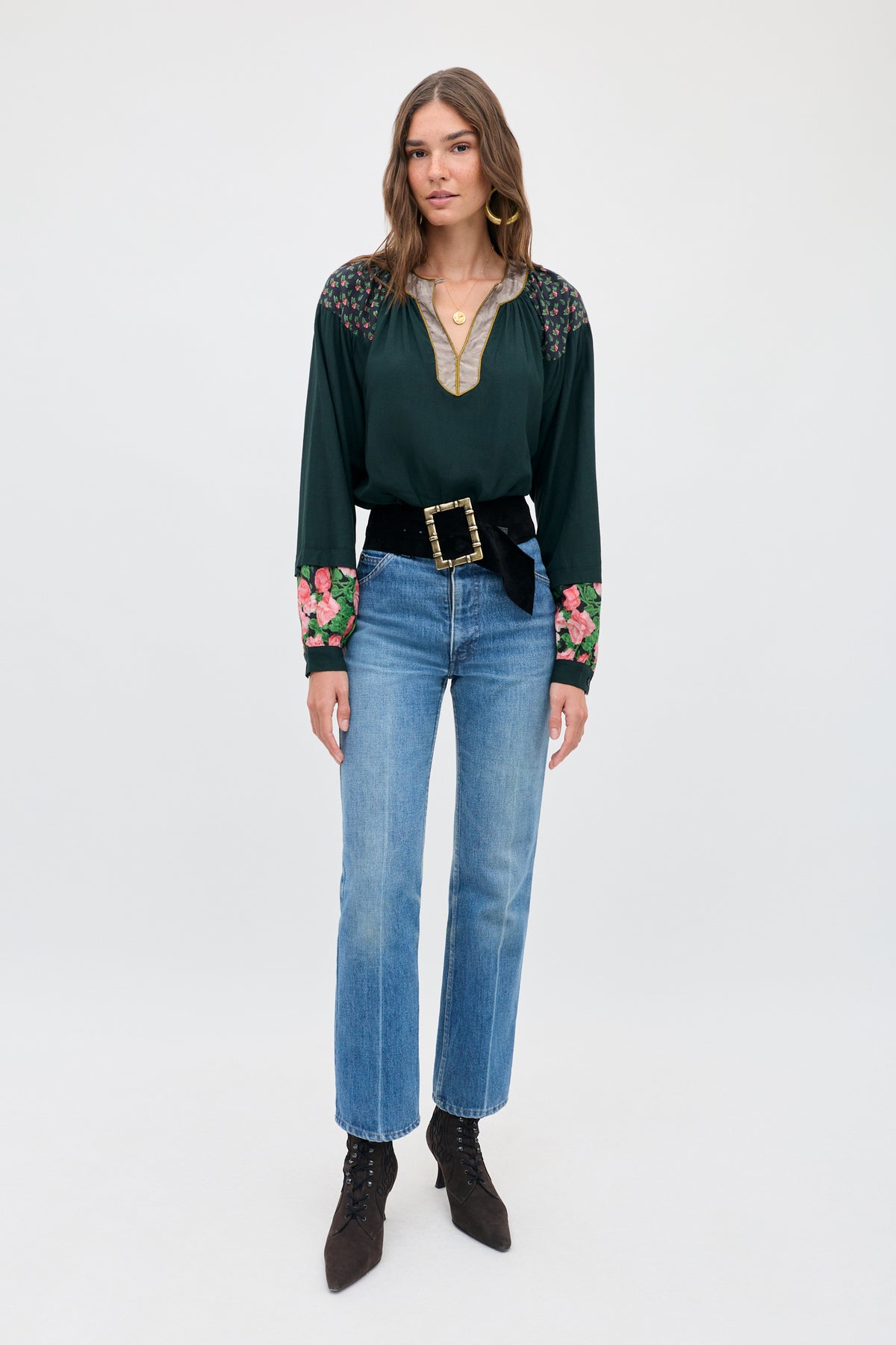 A woman wears a dark green Bria - Rose Mix Pink long-sleeved blouse with floral patterns on the shoulders and cuffs, paired with blue jeans, a black belt with a large gold buckle, and dark pointed-toe lace-up booties.