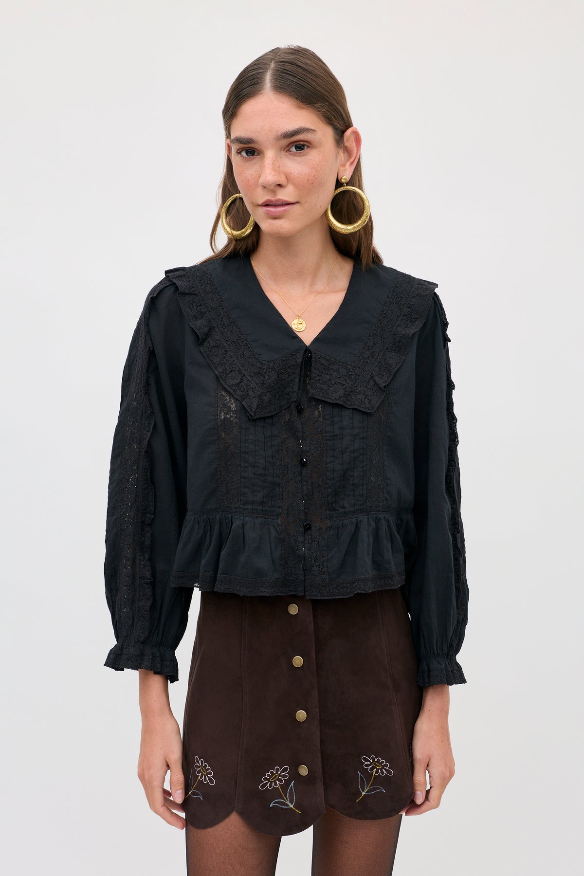 A woman wears a Caris - Shasta Daisy Embroidery Chocolate skirt with small floral embroidery along the scalloped hem, paired with a black long-sleeved blouse featuring a lace-trimmed collar.