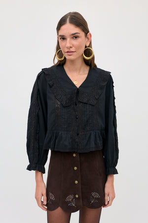 A woman wears a Caris - Shasta Daisy Embroidery Chocolate skirt with small floral embroidery along the scalloped hem, paired with a black long-sleeved blouse featuring a lace-trimmed collar.