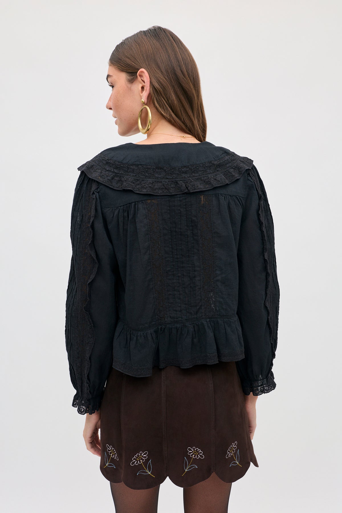 A person wearing a black long-sleeved blouse with a lace collar and the Caris - Shasta Daisy Embroidery Chocolate skirt, which features scalloped edges and daisy embroidery.