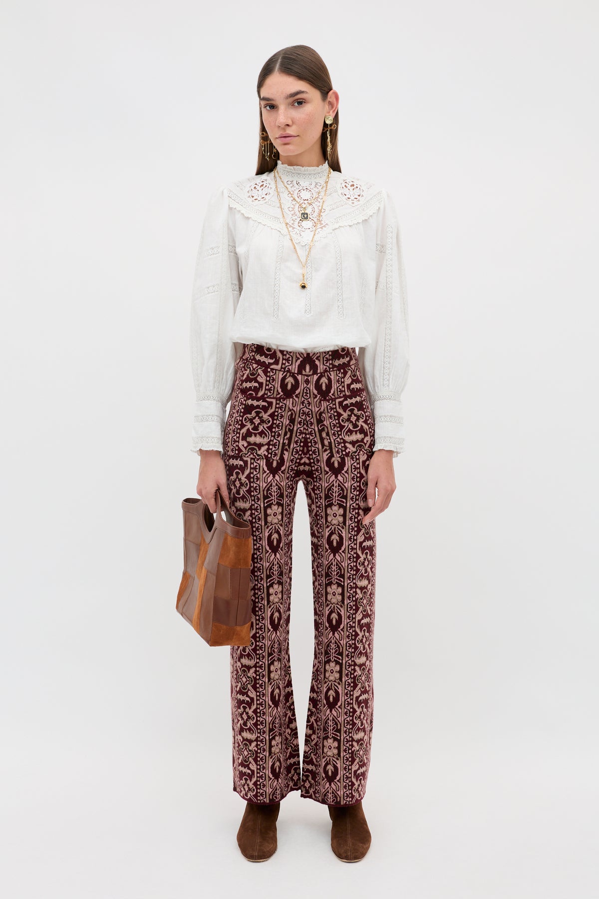 A woman with straight brown hair and a neutral expression stands facing forward against a white background, wearing the Ceara - Tapestry Bloom Bordeaux patterned wide-leg pants, paired with a white lace blouse, brown boots, long gold necklaces, and holding a brown handbag.