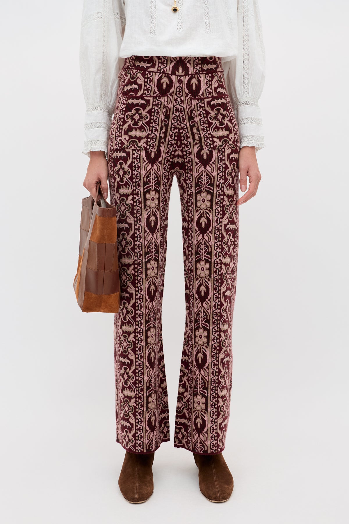 A person is shown from the shoulders down against a plain background, wearing the Ceara - Tapestry Bloom Bordeaux high-waisted patterned pants in maroon and beige, styled with a white blouse, brown boots, and holding a brown and clear rectangular handbag.