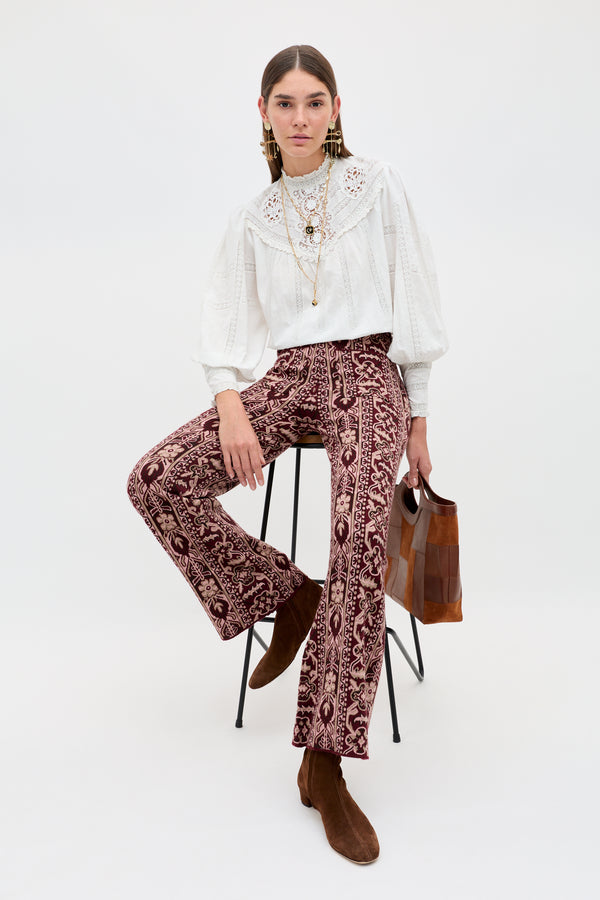 A woman sits on a stool wearing the Ceara - Tapestry Bloom Bordeaux patterned pants, paired with a white lace blouse, brown ankle boots, and holding a brown handbag. Her long dark hair is parted in the middle, and she accessorizes with layered necklaces.