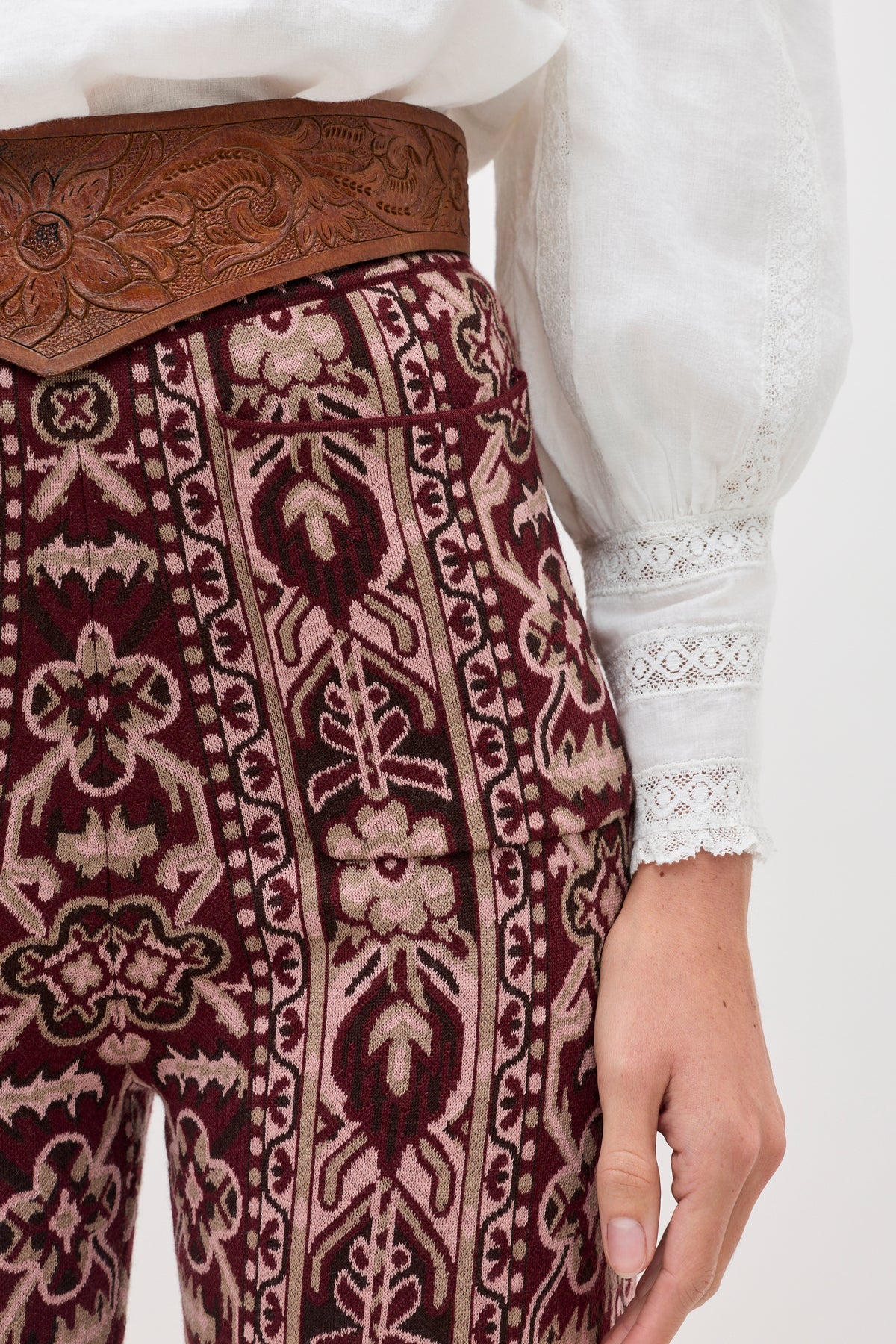 A person wearing the Ceara - Tapestry Bloom Bordeaux patterned maroon pants, paired with a wide brown embossed belt and a white long-sleeve blouse featuring lace details on the sleeves.