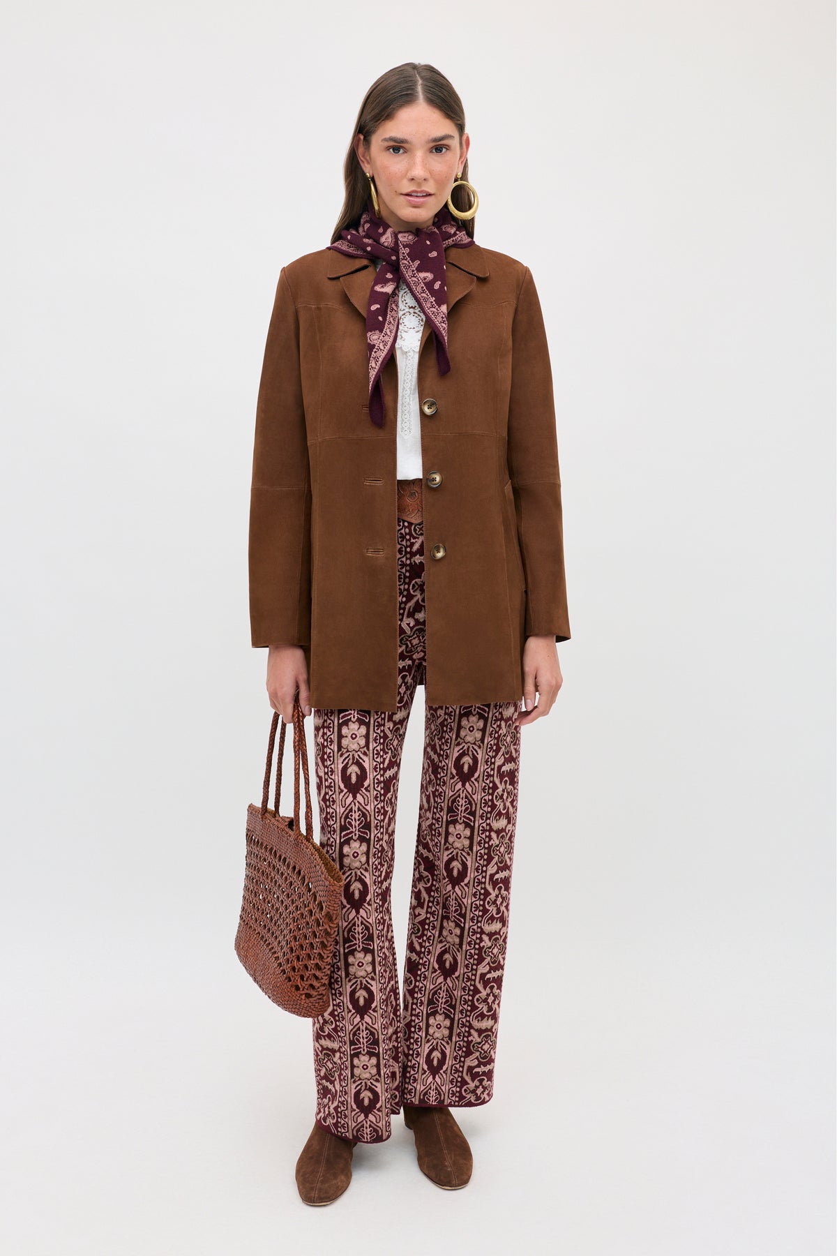 A woman is wearing the Ceara - Tapestry Bloom Bordeaux patterned maroon pants, paired with a brown jacket, white top, matching maroon scarf, hoop earrings, brown shoes, and holding a brown woven bag while standing against a plain light background.