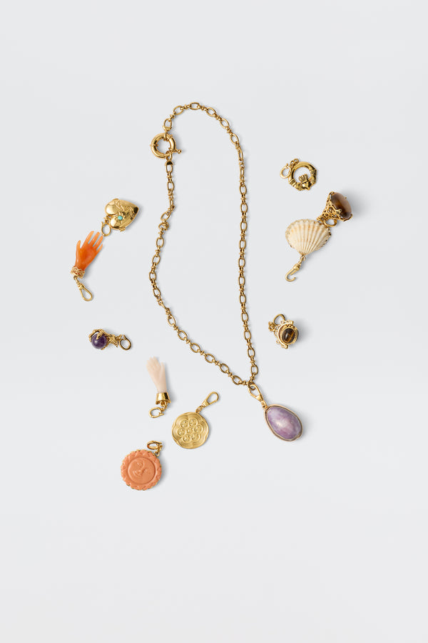 The Chelle - Gold necklace features a gold chain with a purple pendant, surrounded by gold-plated charms and colorful gems—such as a shell, orange hand, coins, and stones—displayed on a white background.