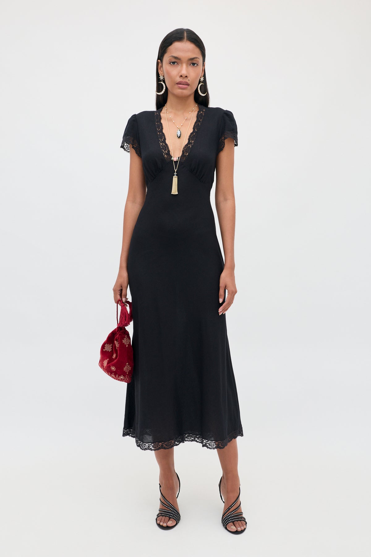 Wearing the Clarice - Black dress, a woman stands against a plain light background, styled with necklaces, black heeled sandals, and a red handbag.