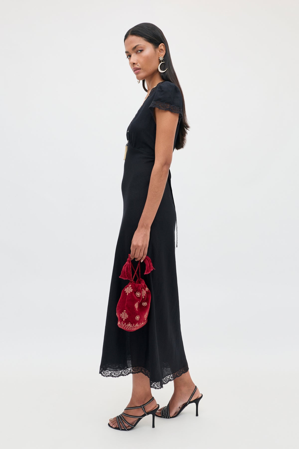 A woman wearing the Clarice - Black midi dress with lace trim holds a small red embroidered bag, paired with black strappy heels and silver earrings, standing against a plain white background, evoking the elegance of a vintage nightdress.