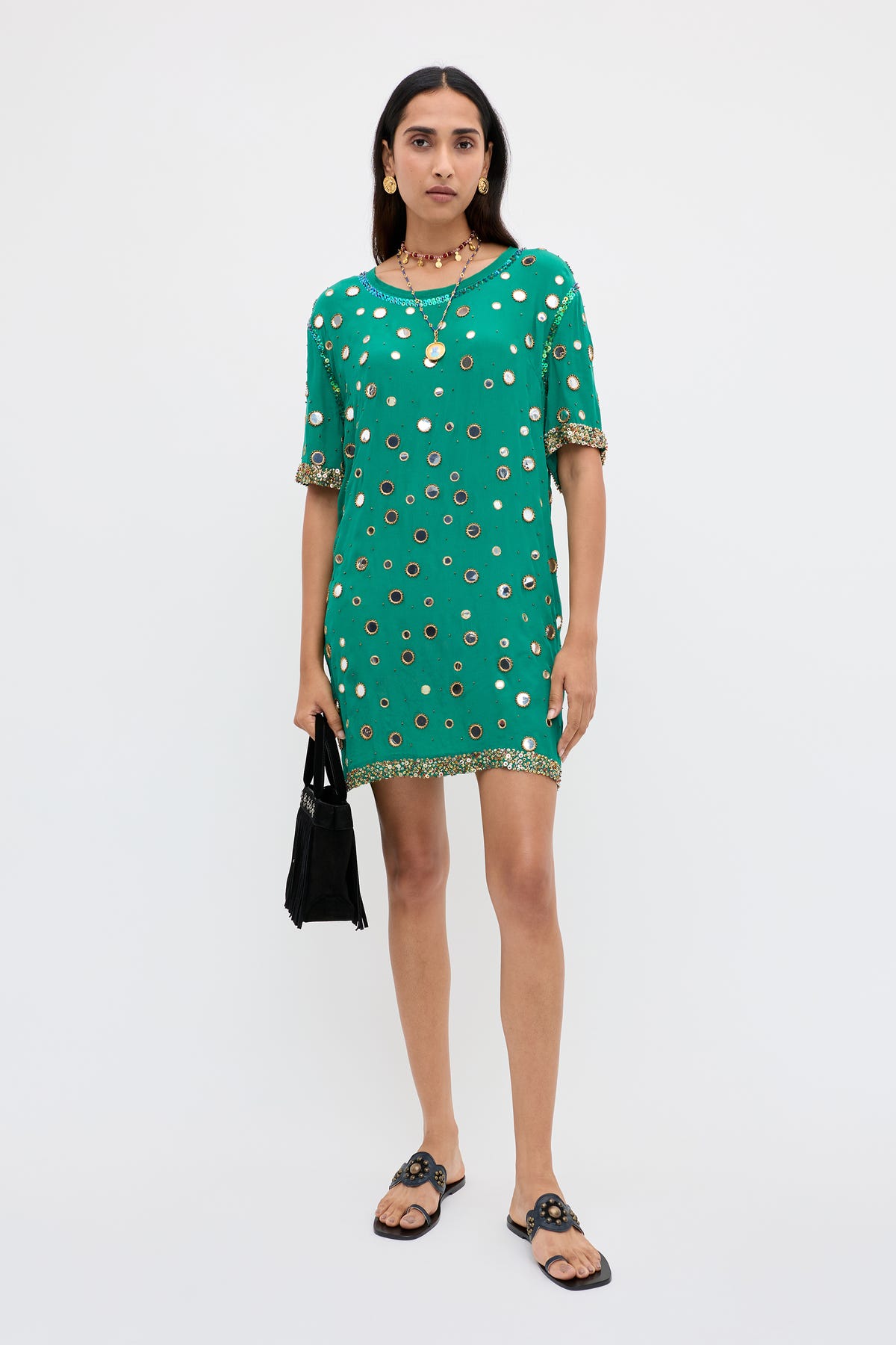 A woman stands against a plain background, wearing the Cruz - Lume Embellishment Dark Green short dress adorned with gold details, paired with black sandals, gold jewelry, and holding a small black handbag.