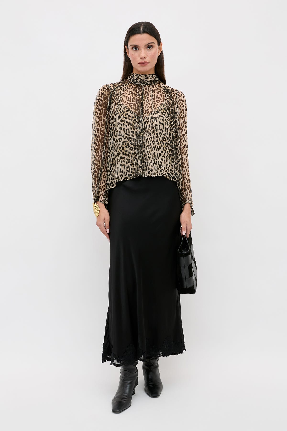 A woman with straight dark hair and a neutral expression stands against a white background wearing a leopard print long-sleeve blouse, a black satin midi skirt, black boots, and carrying the Crystal - Black handbag.