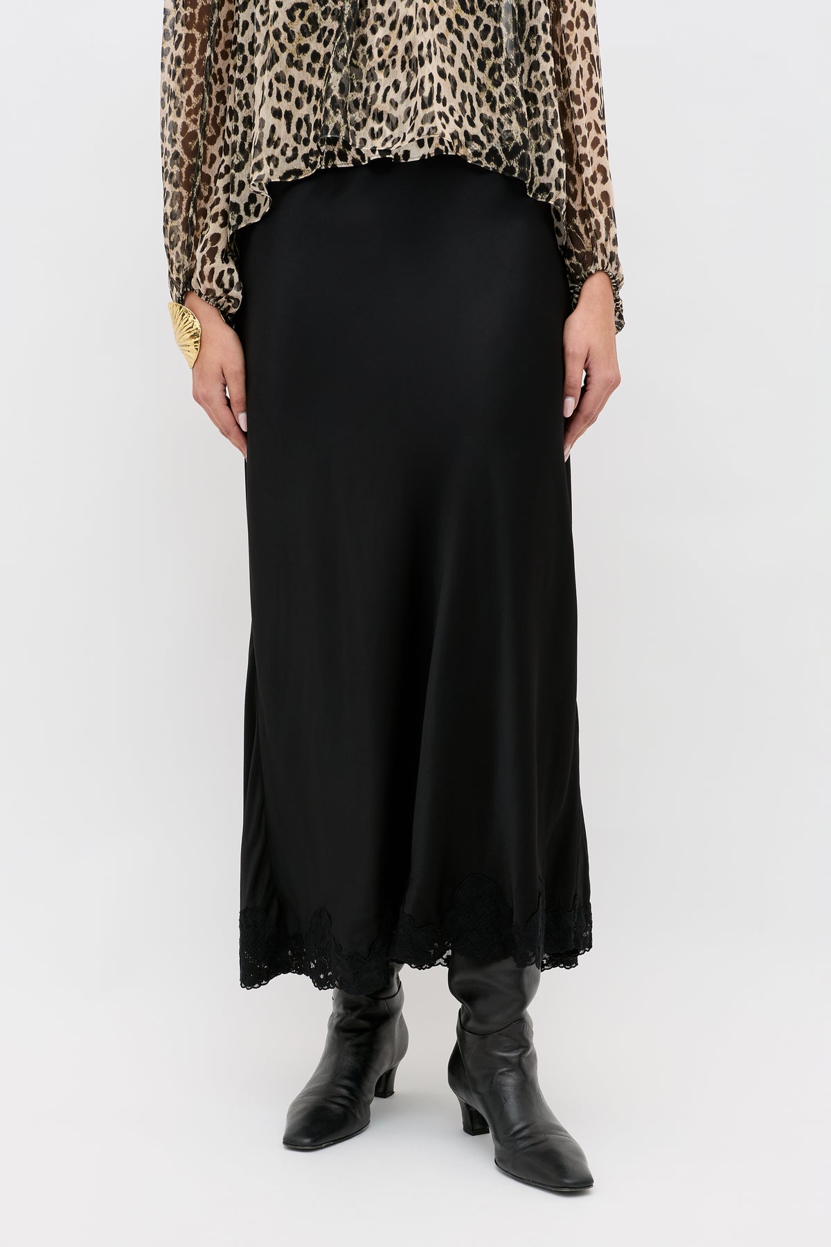 Person wearing a leopard print top, the Crystal - Black bias cut satin midi skirt with lace trim at the hem, black boots, and a large gold ring on their left hand, standing against a plain white background.