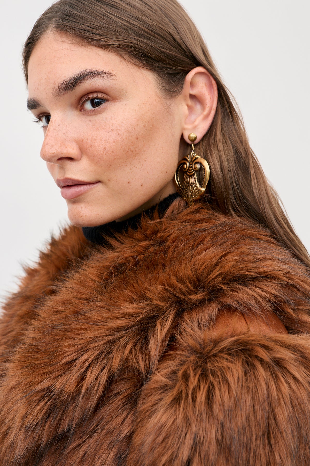 A person wears the Cybil - Brown faux-fur coat and a large, gold-toned metal statement earring.