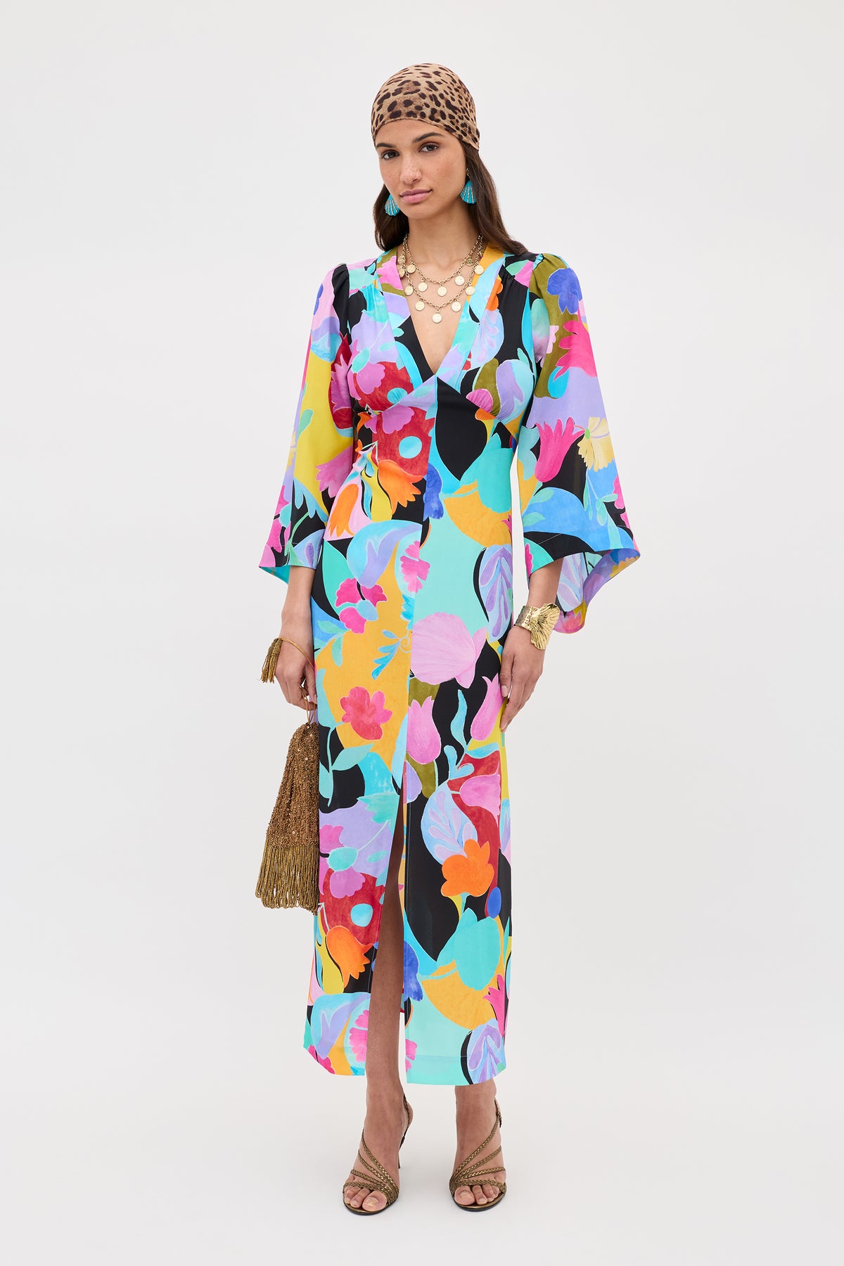 Woman wearing the Dahlia - Abstract Tulip Black dress with dramatic sleeves, a gold necklace, brown animal print headscarf, gold bracelets, tan heeled sandals, and holding a golden fringed handbag.