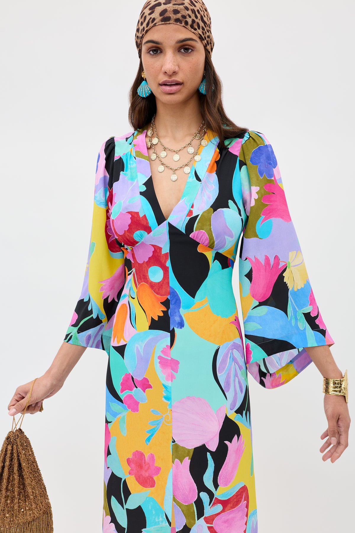 A woman wears the Dahlia - Abstract Tulip Black silk crepe de chine dress with dramatic sleeves, a brown leopard print headscarf, turquoise earrings, gold layered necklaces, and a gold fringed bag against a plain light background.