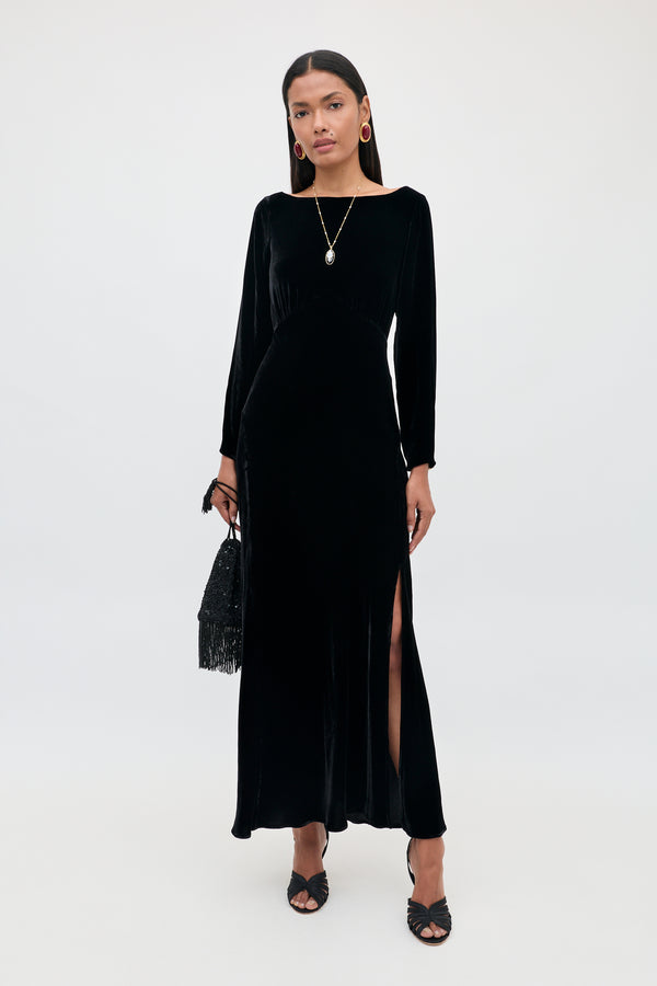 Wearing the Devina - Black gown, a woman stands against a plain background. The long velvet dress features long sleeves and a side slit, highlighting her sophisticated style. She completes the look with black heels, a black handbag, gold hoop earrings, and a pendant necklace.