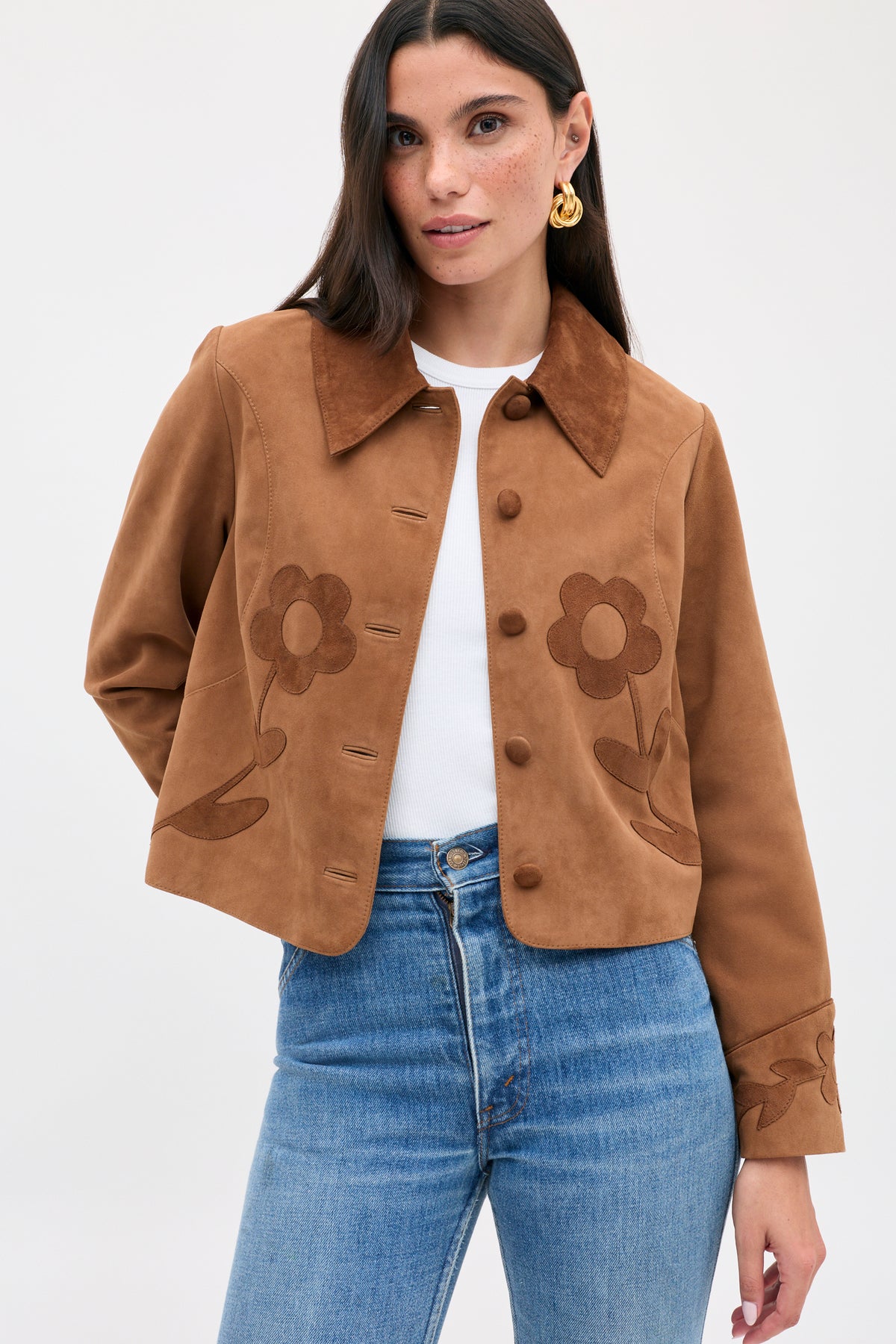 The Dionne - Retro Daisy is a tan suede button-front jacket with a brown collar and dark daisy appliqué on the chest and cuffs, worn over a white shirt and blue jeans.