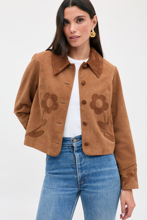 The Dionne - Retro Daisy is a tan suede button-front jacket with a brown collar and dark daisy appliqué on the chest and cuffs, worn over a white shirt and blue jeans.