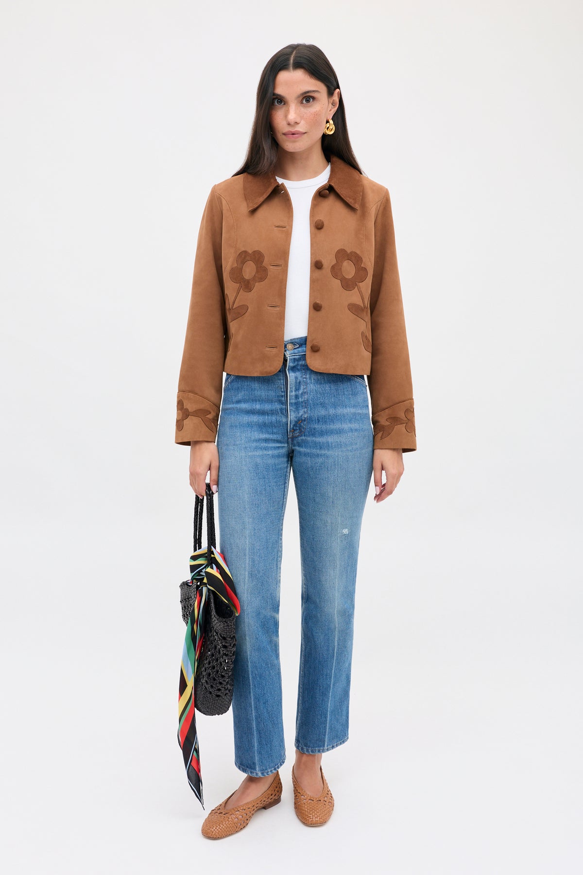 A model wears the Dionne - Retro Daisy brown suede jacket with daisy appliqué over a white shirt and blue jeans, carrying a woven bag with a colorful scarf tied to the handle.