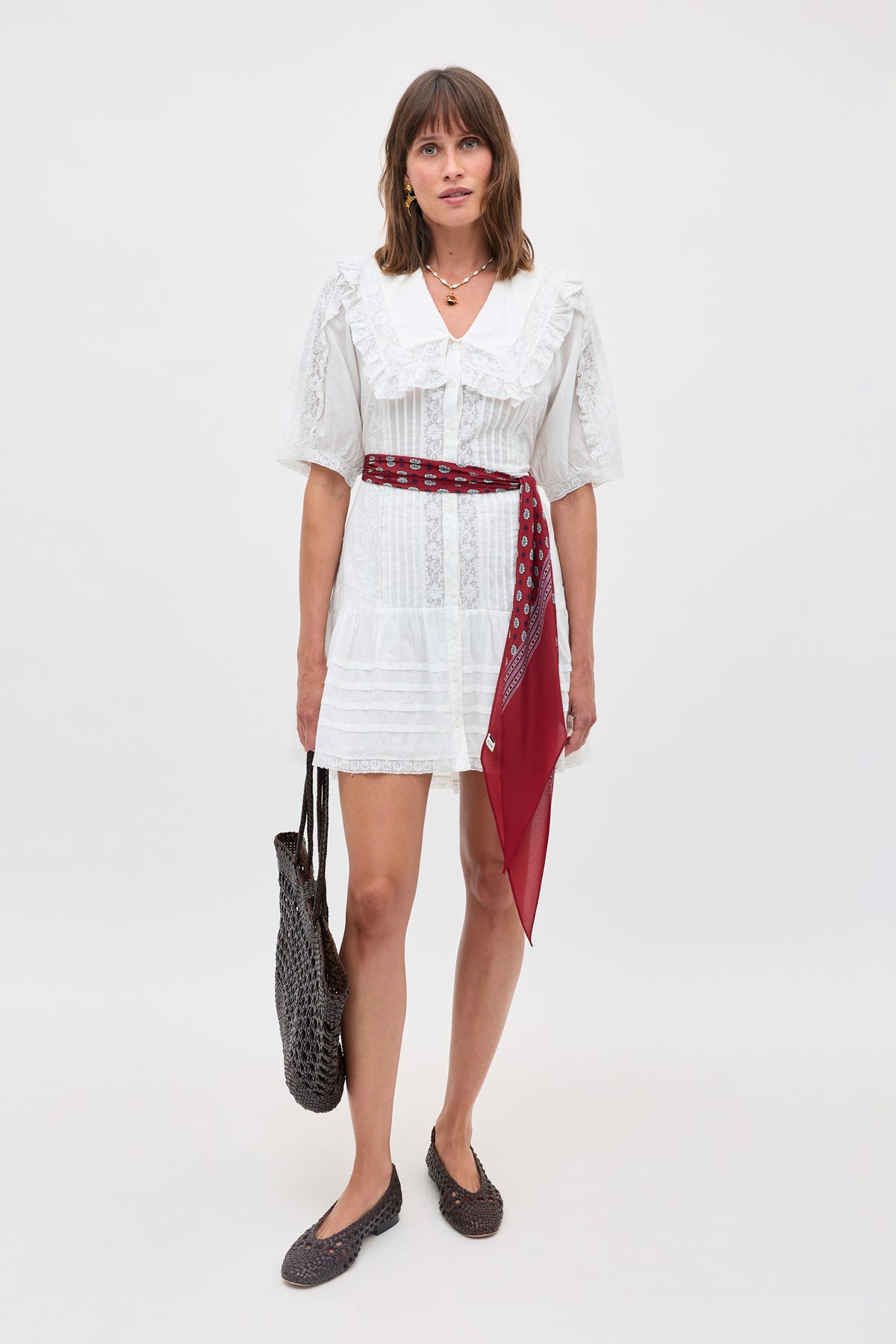 A woman stands against a plain light background wearing the Dove – Pansy Embroidery White short cotton-voile lace dress with puff sleeves, styled with a red patterned scarf as a belt, brown woven flats, and holding a dark brown woven bag.