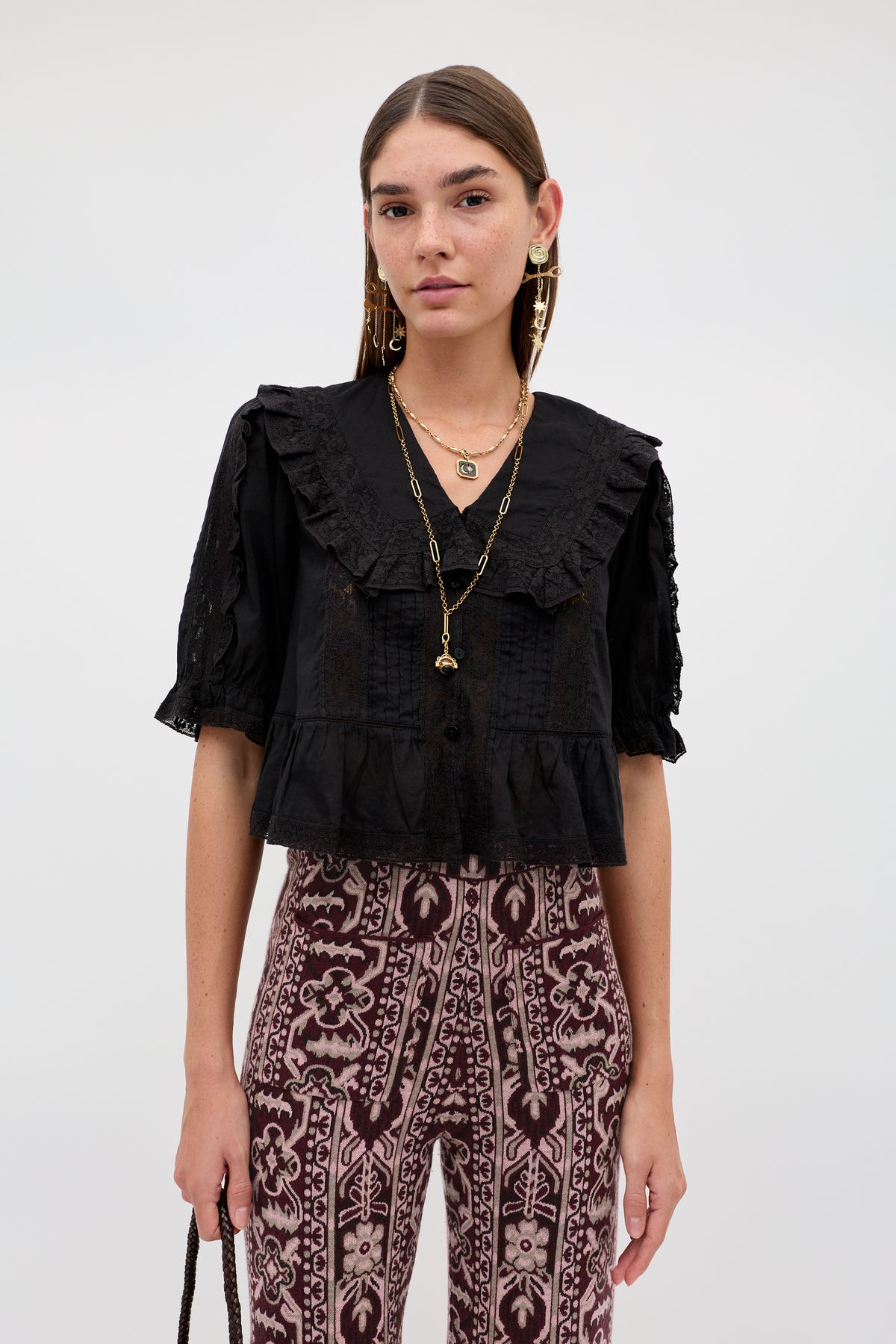Wearing the “Ellery - Black” ruffled collar blouse with lace details, the woman pairs it with patterned high-waisted pants, layered gold necklaces, earrings, and holds a black purse while standing against a plain light background.