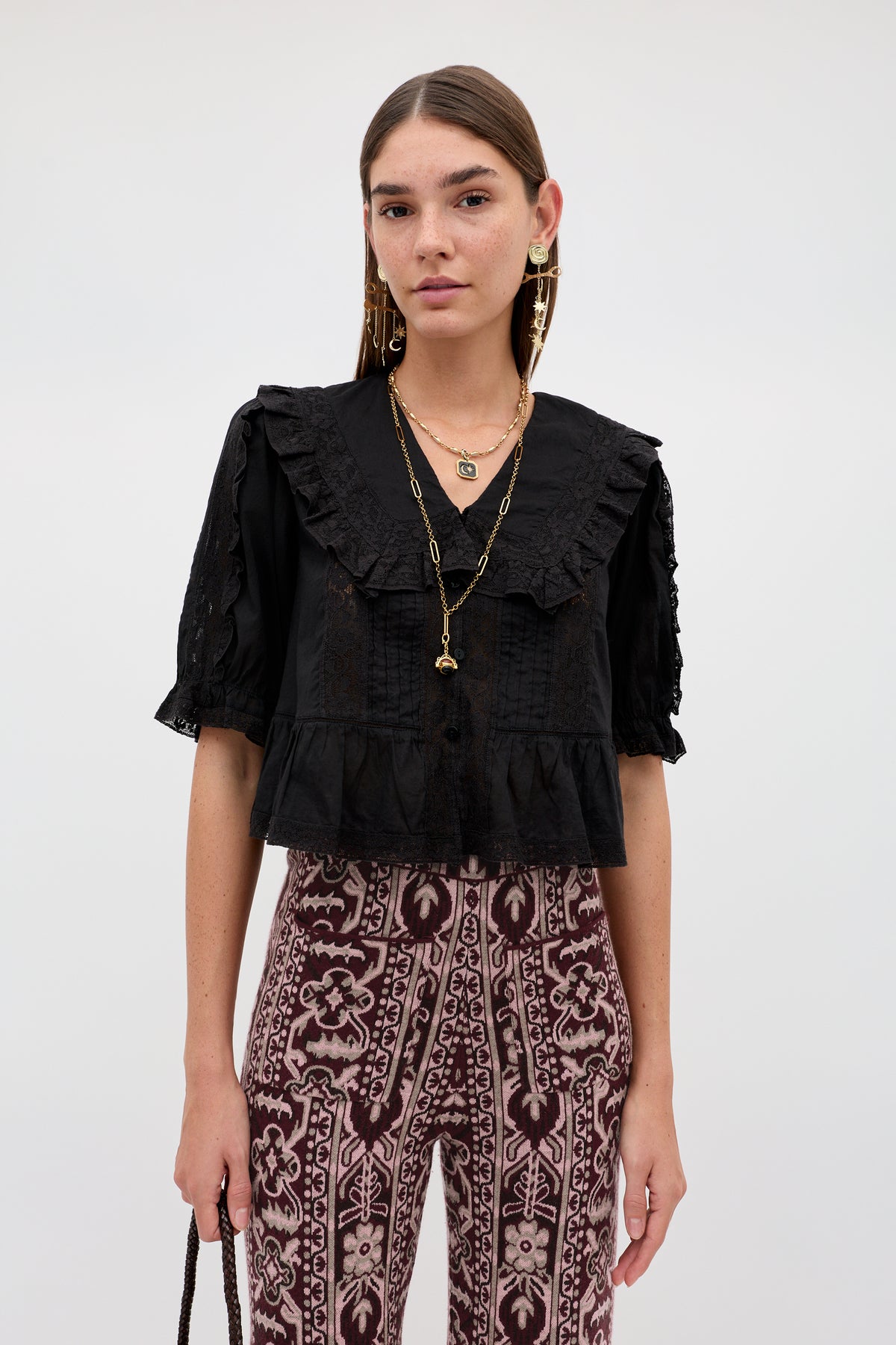Wearing the “Ellery - Black” ruffled collar blouse with lace details, the woman pairs it with patterned high-waisted pants, layered gold necklaces, earrings, and holds a black purse while standing against a plain light background.