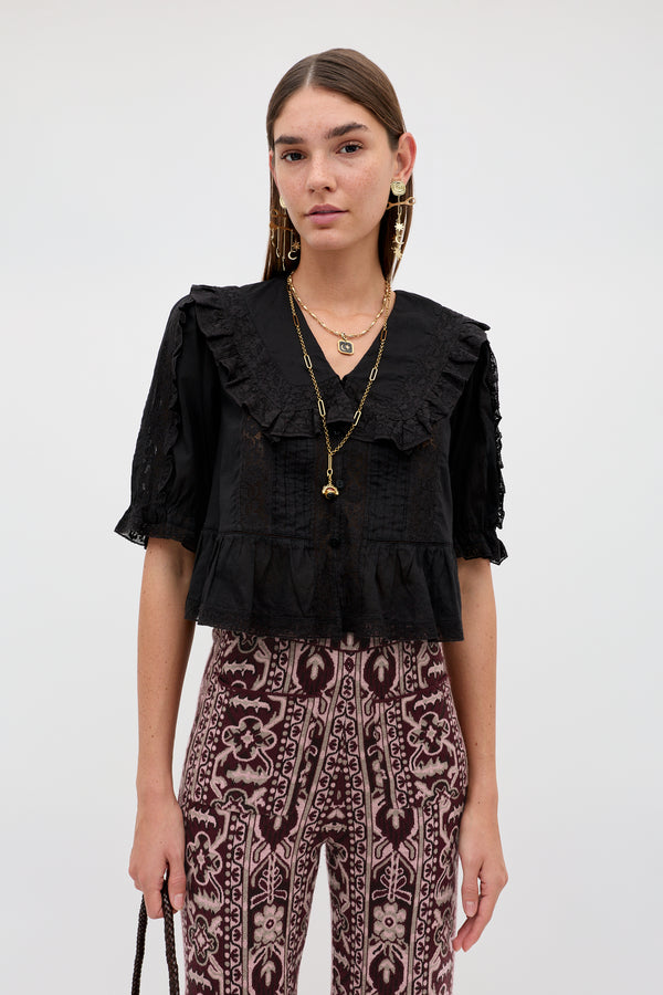Wearing the “Ellery - Black” ruffled collar blouse with lace details, the woman pairs it with patterned high-waisted pants, layered gold necklaces, earrings, and holds a black purse while standing against a plain light background.