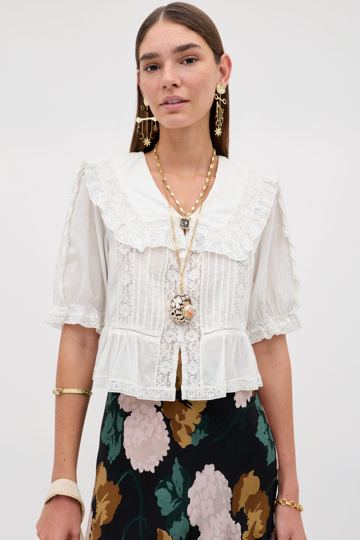 Wearing the Ellery - White blouse, a woman stands against a plain light background. The vintage-inspired top, crafted from airy cotton voile with delicate lace details and a ruffled collar, beautifully complements her long brown hair. She pairs it with gold jewelry and a floral skirt for an elegant look.