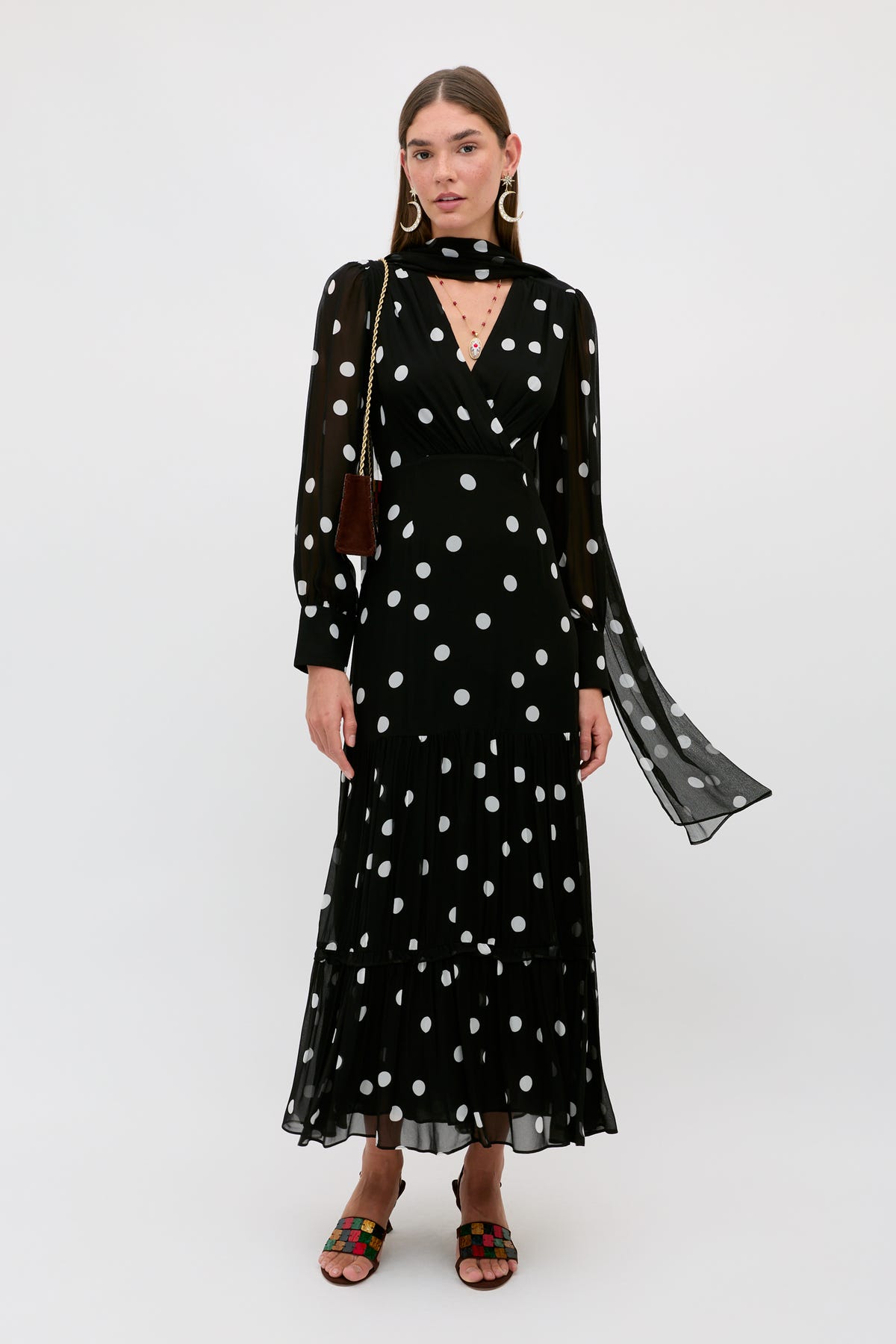 A woman stands against a plain white background wearing the “Elsie - Cosmos Spot Black” dress, a long, blousy polka dot design with sheer long sleeves and a matching scarf, paired with colorful sandals.