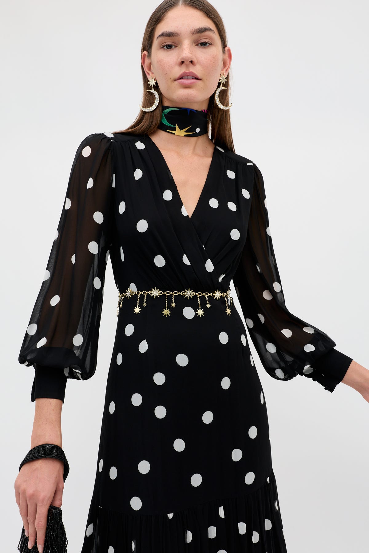 A woman wears the Elsie - Cosmos Spot Black dress with chic polka dots and sheer long sleeves, styled with a gold star belt, star earrings, and a black floral neck scarf, standing against a plain light background.