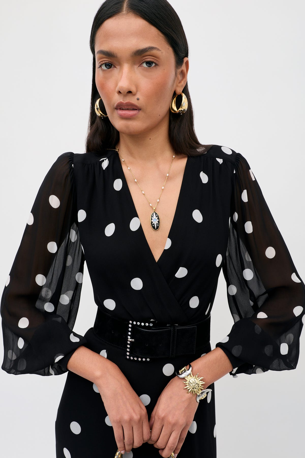 A woman stands against a plain background with her hands clasped, wearing the Elsie - Cosmos Spot Black dress—a blousy piece adorned with classic white polka dots for a timeless look. She completes her outfit with sheer sleeves, gold earrings, a cameo necklace, and a decorative bracelet.