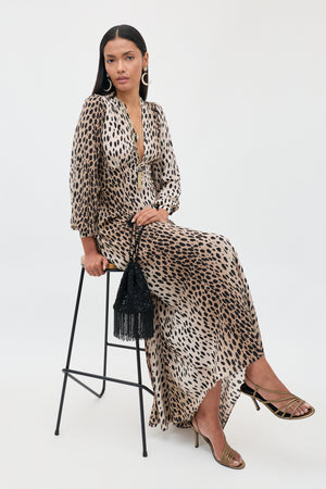 Seated gracefully on a tall stool, a woman wears the Emory - Antelope Spot Black dress, an elegant leopard-print design in luxurious silk crepe de chine with a plunging neckline. She completes her special occasion ensemble with gold sandals and statement earrings, holding a black fringed handbag. Her long, straight hair flows beautifully as she poses against a simple light backdrop.