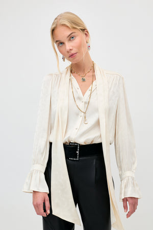 Wearing the Evanora - Floral Trail blouse, featuring a cream hue with ruffled cuffs and a long necktie detail, the woman poses against a plain light background. She completes her look with layered gold necklaces, statement drop earrings, black pants accented by a studded belt.