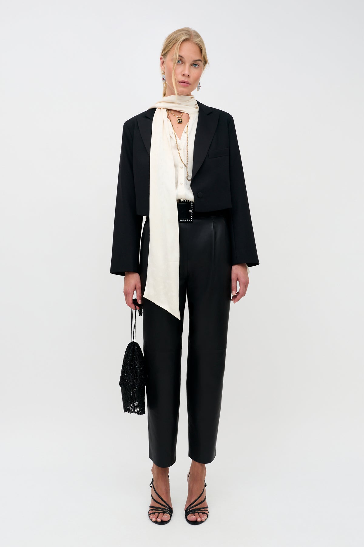 Woman standing against a white background, wearing the Evanora - Floral Trail blazer and matching high-waisted pants in black, a cream blouse with a long scarf, black strappy heels, and holding a small black handbag.
