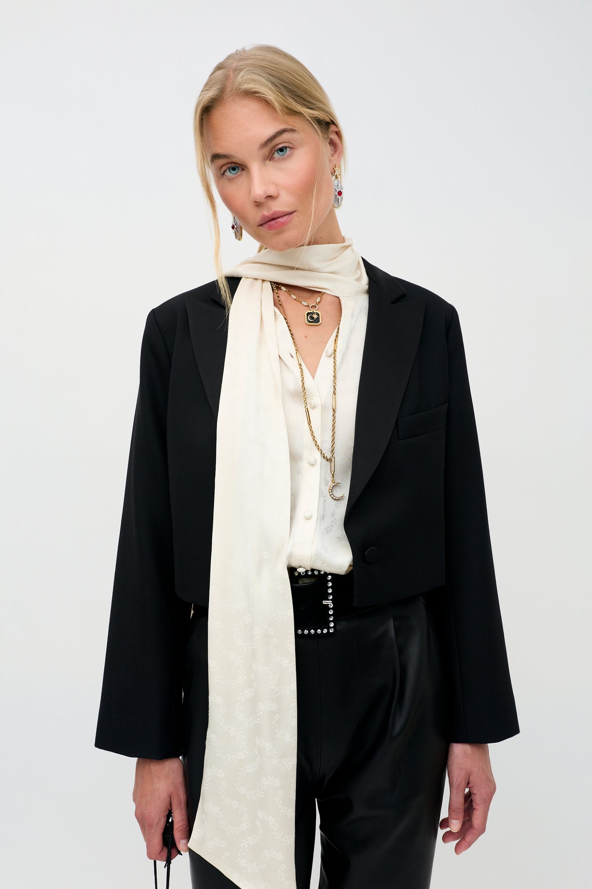 Wearing the Evanora - Floral Trail, a woman with blonde hair poses against a plain light background. She styles it with a black blazer, white blouse, long white scarf, black pants, layered necklaces, and earrings.