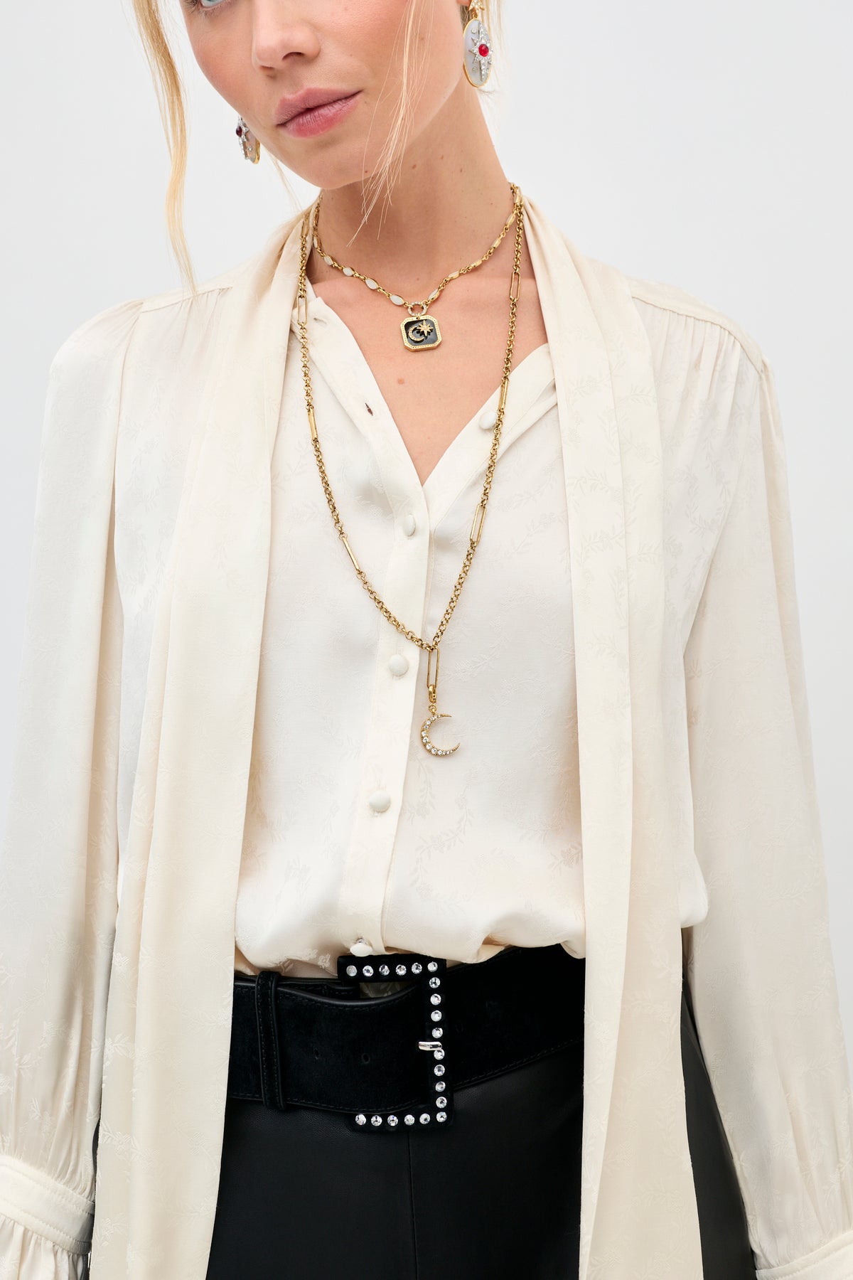 A woman wears the Eta - Gold necklace layered over a cream blouse, paired with statement earrings and a black belt with silver studs. Her face is partially visible, and her blonde hair is styled in a loose, casual look.
