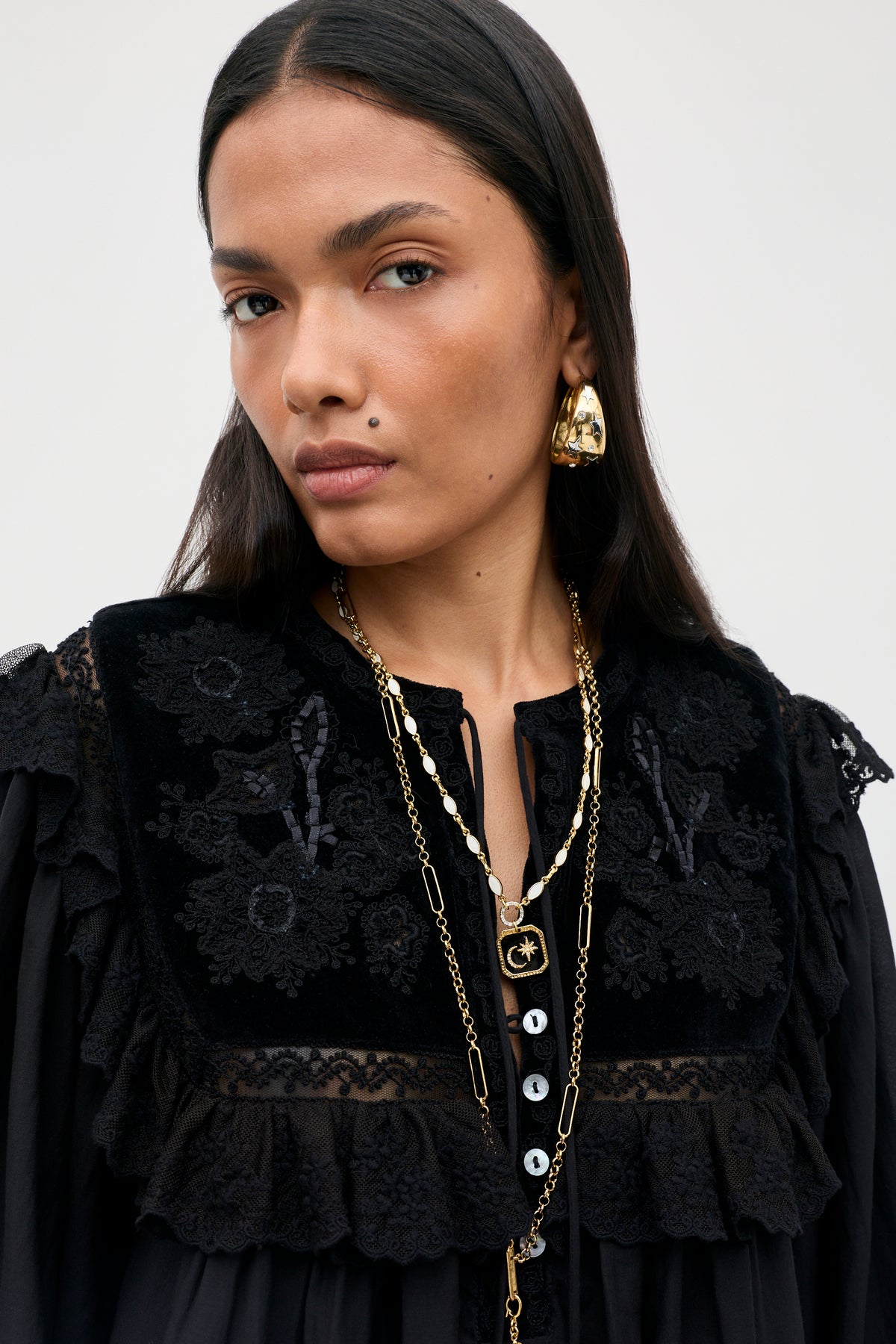 A woman with long dark hair wears the Inez - Lilly Embroidery Black dress, paired with gold earrings and layered gold charm necklaces, gazing confidently at the camera against a neutral background.