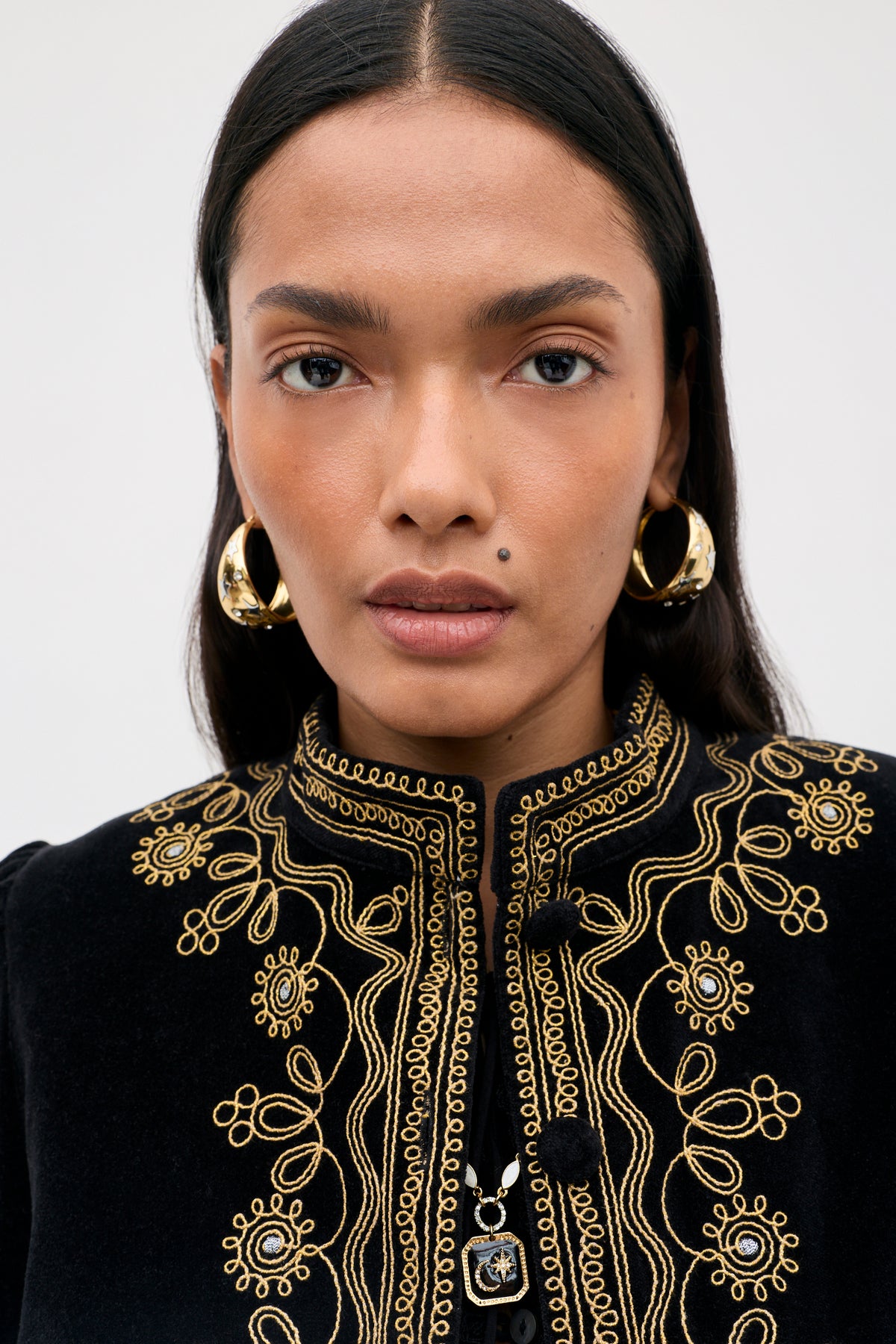 A person with long dark hair, wearing gold hoop earrings and a black top with intricate gold embroidery, looks directly at the camera with a neutral expression. The luxurious feel of the ensemble is elevated by the elegant Denver - Soutache Black velvet jacket.