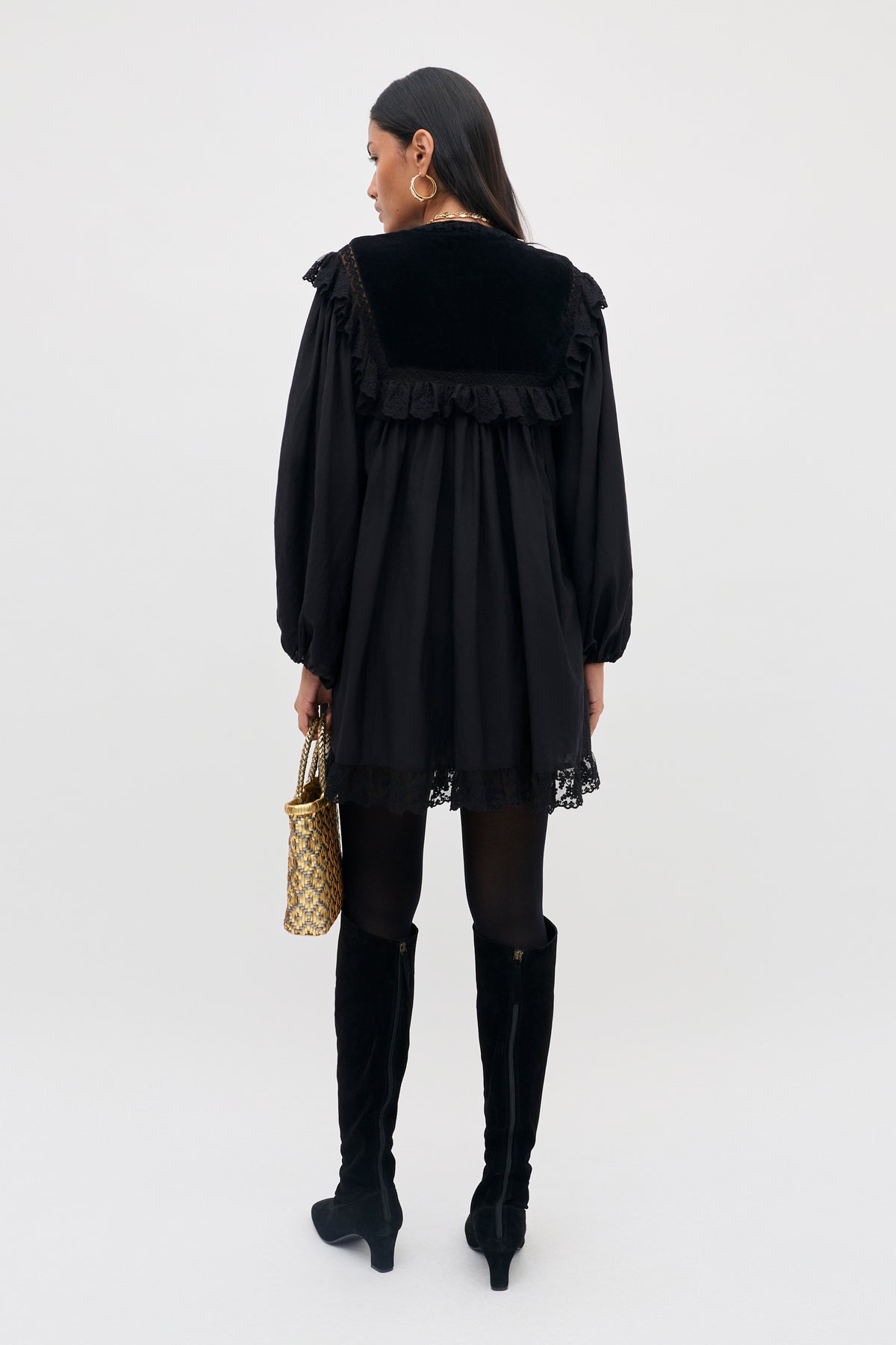 A person with long dark hair is photographed from the back against a plain background, wearing the Inez - Lilly Embroidery Black dress, styled with black tights, knee-high black boots, gold hoop earrings, and holding a patterned gold handbag.
