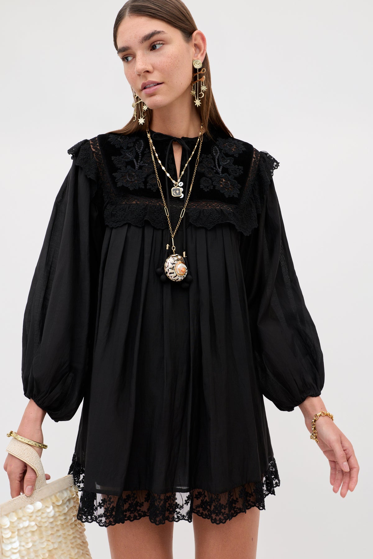 A model wears the Inez - Lilly Embroidery Black, a black long-sleeved mini dress with a pleated bodice, lace trim, and floral embroidery on the yoke, paired with layered necklaces, statement earrings, and a small shell-embellished bag.