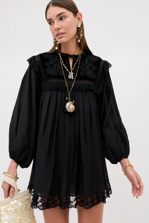 A model wears the Inez - Lilly Embroidery Black, a black long-sleeved mini dress with a pleated bodice, lace trim, and floral embroidery on the yoke, paired with layered necklaces, statement earrings, and a small shell-embellished bag.